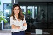 © insta_photos - Smiling confident Latin professional mid aged business woman corporate manager leader, happy beautiful 40s mature female executive standing at office window arms crossed looking at camera, portrait.