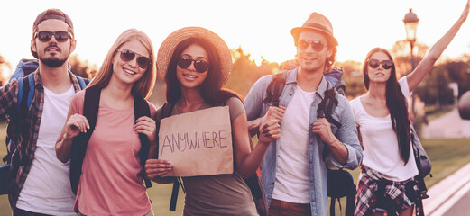 Wall Mural - Group of young people with backpacks hitchhiking on the road and looking happy