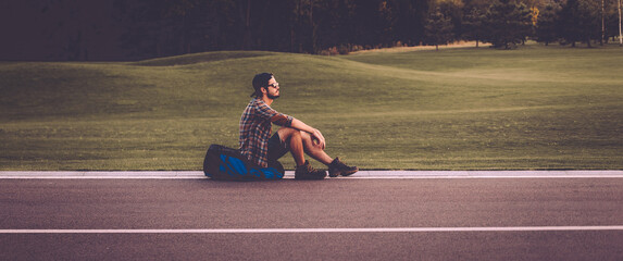 Canvas Print - Side view of handsome young man sitting on his backpack at the side of the road