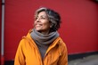 © Leon Waltz - Portrait of a smiling middle-aged woman in a yellow coat and gray scarf against a red wall