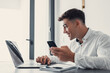 © Daniel - Confident man sitting at desk taking break in work with electronic documents on laptop to make answer telephone call. Smiling young guy freelancer synchronize data between home computer and smartphone