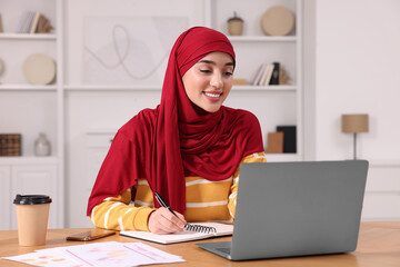 Wall Mural - Muslim woman writing notes near laptop at wooden table at home