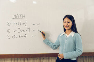 Asian female teacher is teaching students at the classroom while pointing at numbers on the white board.