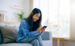© Monster Ztudio - Happy young asian woman relax on comfortable couch at home, typing chat message on smartphone, smiling girl use cell phone chatting, searching information on browser wireless internet, online shopping