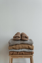 Wall Mural - Stack of warm neutral beige clothes on wooden stool over white wall. Fluffy slippers, woolen pullovers and sweaters