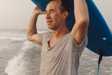 Portrait Of Mature Surfer Looking Ahead While Carrying Blue Surfboard On Head With Hands, Standing On Wavy Seaside.