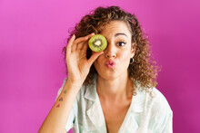Woman Puckering And Holding Slice Of Kiwi Over Eye Against Magenta Background