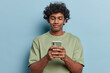© WHstudio Leushin N - People positive emotions concept. Indoor shot of young happy Hindu male wearing olive t shirt standing in centre isolated on blue background holding smartphone reading message with good news