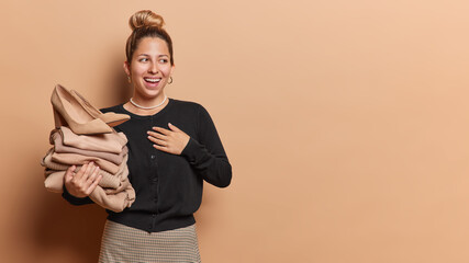 Wall Mural - Joyful European woman keeps hand on chest laughs happily and looks aside holds stack of brown folded clothes and high heeled shoes wears black jumper skirt isolated over beige background copy space