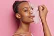 Beautiful woman with curly combed hair puts on facial mask for skin treatment smiles gently wears t shirt isolated over pink wall. Young female model removing from face skincare hydrating sheet mask