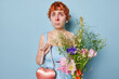 Photo of redhead young European woman has red watery eyes holds small heart shaped bag and bouquet of wild flowers suffers from allergy to pollen comes on date isolated over blue background.