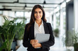 © AI_images - Smiling latin young professional business woman corporate marketing manager, female worker holding digital tablet computer fintech tab at work standing in modern company office looking at camera.