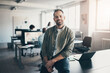 © Flamingo Images - Smiling businessman leaning on an office desk with colleagues in the background