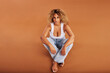 © Flamingo Images - Confident woman wearing jeans and a tank top sitting on an orange background