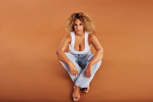 Confident Woman Wearing Jeans And A Tank Top Sitting On An Orange Background