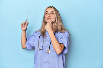 Young caucasian nurse in blue studio looking sideways with doubtful and skeptical expression.