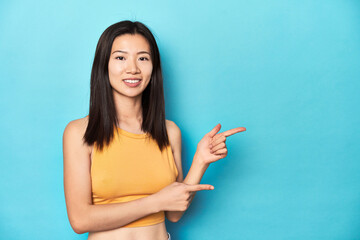 Wall Mural - Asian woman in summer yellow top, studio setup, excited pointing with forefingers away.
