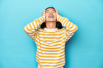 Wall Mural - Asian woman in striped yellow sweater, laughs joyfully keeping hands on head. Happiness concept.