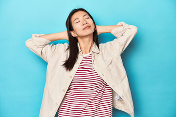 Wall Mural - Asian woman in layered shirt and striped t-shirt, feeling confident, with hands behind the head.