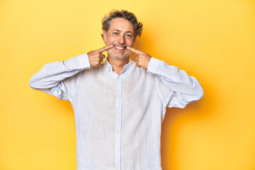 Wall Mural - Middle-aged man posing on a yellow backdrop smiles, pointing fingers at mouth.