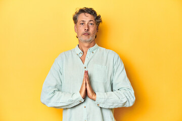 Wall Mural - Middle-aged man posing on a yellow backdrop praying, showing devotion, religious person looking for divine inspiration.