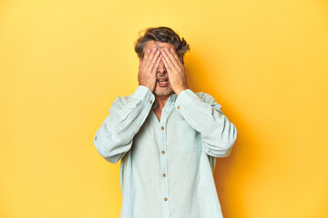 Wall Mural - Middle-aged man posing on a yellow backdrop afraid covering eyes with hands.