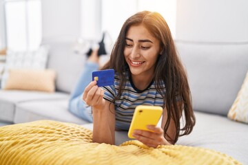 Wall Mural - Young hispanic girl using smartphone and credit card lying on sofa at home