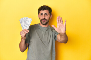 Caucasian man holding dollars, yellow studio shot standing with outstretched hand showing stop sign, preventing you.