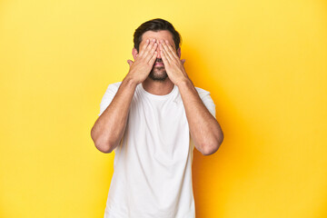 Caucasian man in white t-shirt on yellow studio background afraid covering eyes with hands.