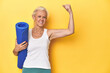 Fitness woman with yoga mat, sportswear on yellow backdrop raising fist after a victory, winner concept.