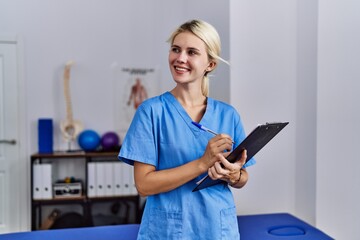 Poster - Young blonde woman pysiotherapist smiling confident writing on document at rehab clinic