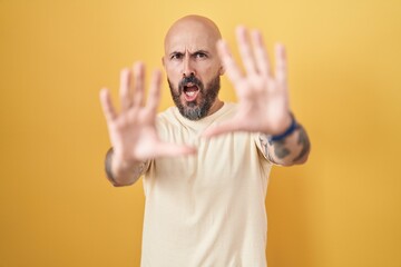 Poster - Hispanic man with tattoos standing over yellow background afraid and terrified with fear expression stop gesture with hands, shouting in shock. panic concept.