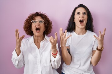 Poster - Hispanic mother and daughter wearing casual white t shirt over pink background crazy and mad shouting and yelling with aggressive expression and arms raised. frustration concept.