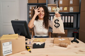 Sticker - Young hispanic woman working at small business ecommerce holding money bag smiling happy doing ok sign with hand on eye looking through fingers