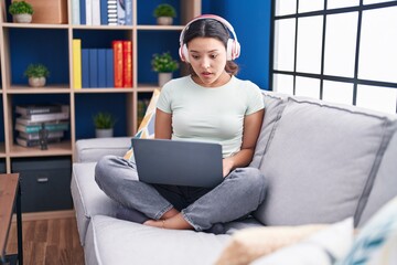 Sticker - Hispanic young woman using laptop at home wearing headphones scared and amazed with open mouth for surprise, disbelief face