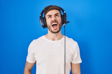 Poster - Hispanic man with beard listening to music wearing headphones angry and mad screaming frustrated and furious, shouting with anger. rage and aggressive concept.