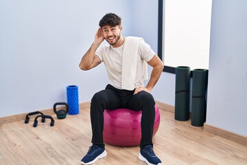 Canvas Print - Hispanic man with beard sitting on pilate balls at yoga room smiling with hand over ear listening an hearing to rumor or gossip. deafness concept.