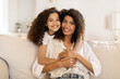 © Home-stock - Happy latin girl hugging her beautiful mother and smiling together at camera, sitting on sofa in living room, adorable family embracing at home