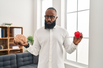 Poster - African american man working at therapy office holding brain and heart skeptic and nervous, frowning upset because of problem. negative person.