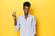 © Asier - African businesswoman, blue striped shirt, yellow backdrop, joyful and carefree showing a peace symbol with fingers.