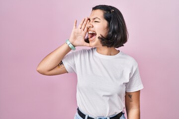 Poster - Young hispanic woman wearing casual white t shirt over pink background shouting and screaming loud to side with hand on mouth. communication concept.