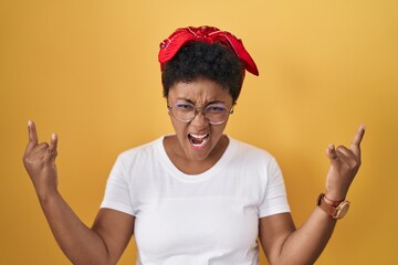 Sticker - Young african american woman standing over yellow background shouting with crazy expression doing rock symbol with hands up. music star. heavy music concept.