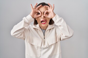 Wall Mural - Middle age hispanic woman standing over isolated background doing ok gesture like binoculars sticking tongue out, eyes looking through fingers. crazy expression.
