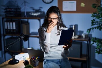 Canvas Print - Young brazilian woman using touchpad at night working at the office smelling something stinky and disgusting, intolerable smell, holding breath with fingers on nose. bad smell