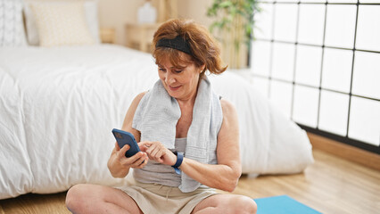 Poster - Middle age woman sitting on yoga mat using smartphone at bedroom