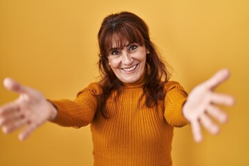 Poster - Middle age hispanic woman standing over yellow background looking at the camera smiling with open arms for hug. cheerful expression embracing happiness.