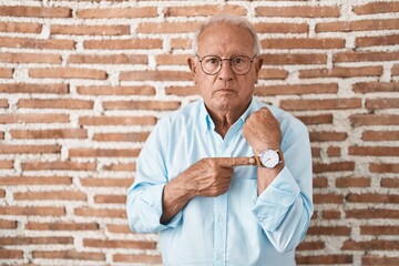 Canvas Print - Senior man with grey hair standing over bricks wall in hurry pointing to watch time, impatience, looking at the camera with relaxed expression