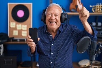 Wall Mural - Senior man with grey hair showing smartphone screen at music studio celebrating victory with happy smile and winner expression with raised hands