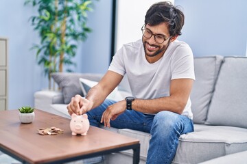 Wall Mural - Young hispanic man inserting coin on piggy bank at home