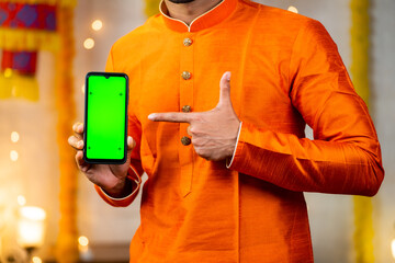 Wall Mural - Close up shot of indian man in traditional ethnic wear showing green screen mobile phone by pointing finger by looking camera - concept of app or application, promotion, festival sales and offers